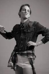 Monochrome portrait of a young male model wearing a denim jacket, white shorts, and a plaid shirt tied around his waist.