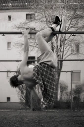 Monochrome image of a person hanging upside down from a metal bar wearing a plaid skirt and sneakers.