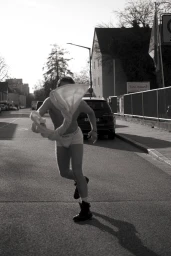 A person running down a street in daylight holding up a white fabric garment, wearing underwear and boots.