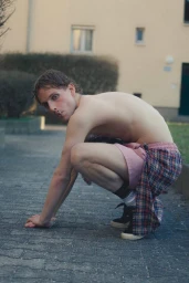 Shirtless young man crouching on a paved walkway wearing pink shorts and a plaid shirt tied around his waist