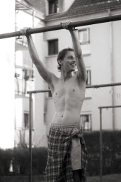 Shirtless man with plaid fabric tied around his waist hanging from an outdoor bar against a residential building.