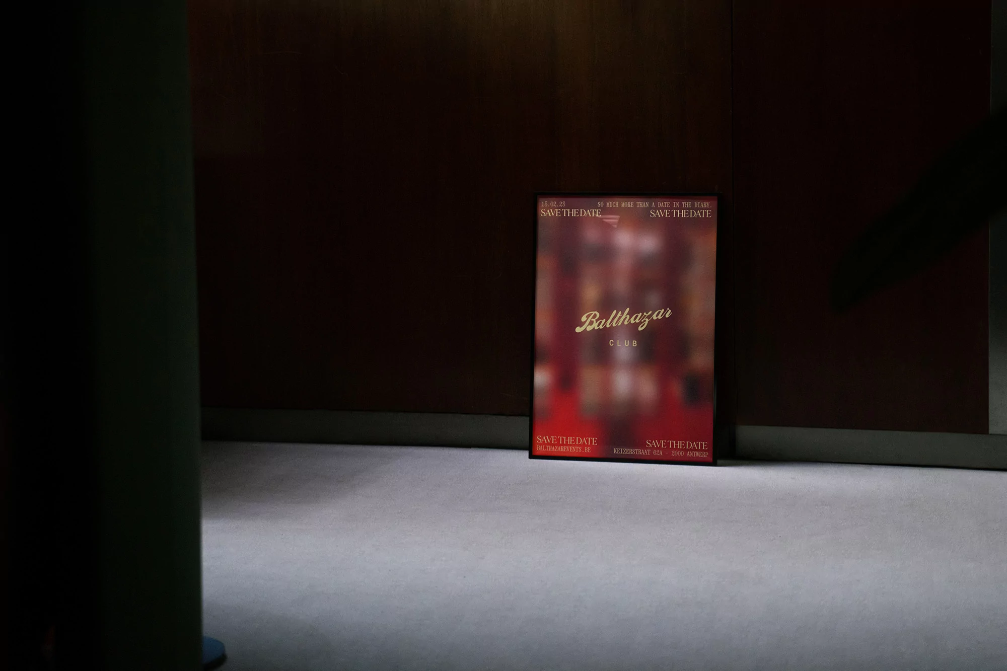 A blurred promotional poster for the Balthazar Club leans against a dark paneled wall, bathed in soft, indirect lighting.