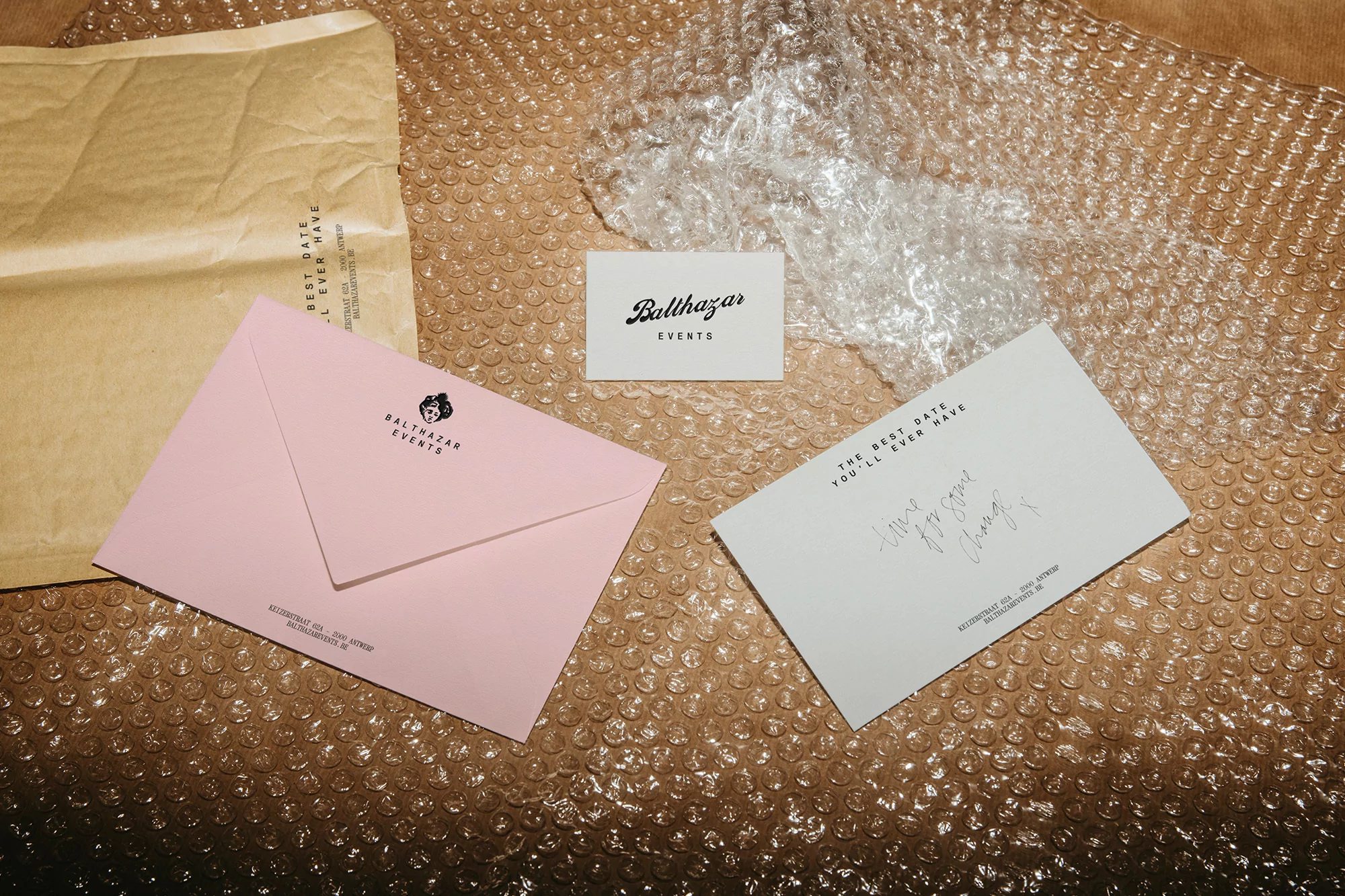 Image of event stationary featuring a pink envelope, a business card, and a white greeting card on tan bubble wrap.