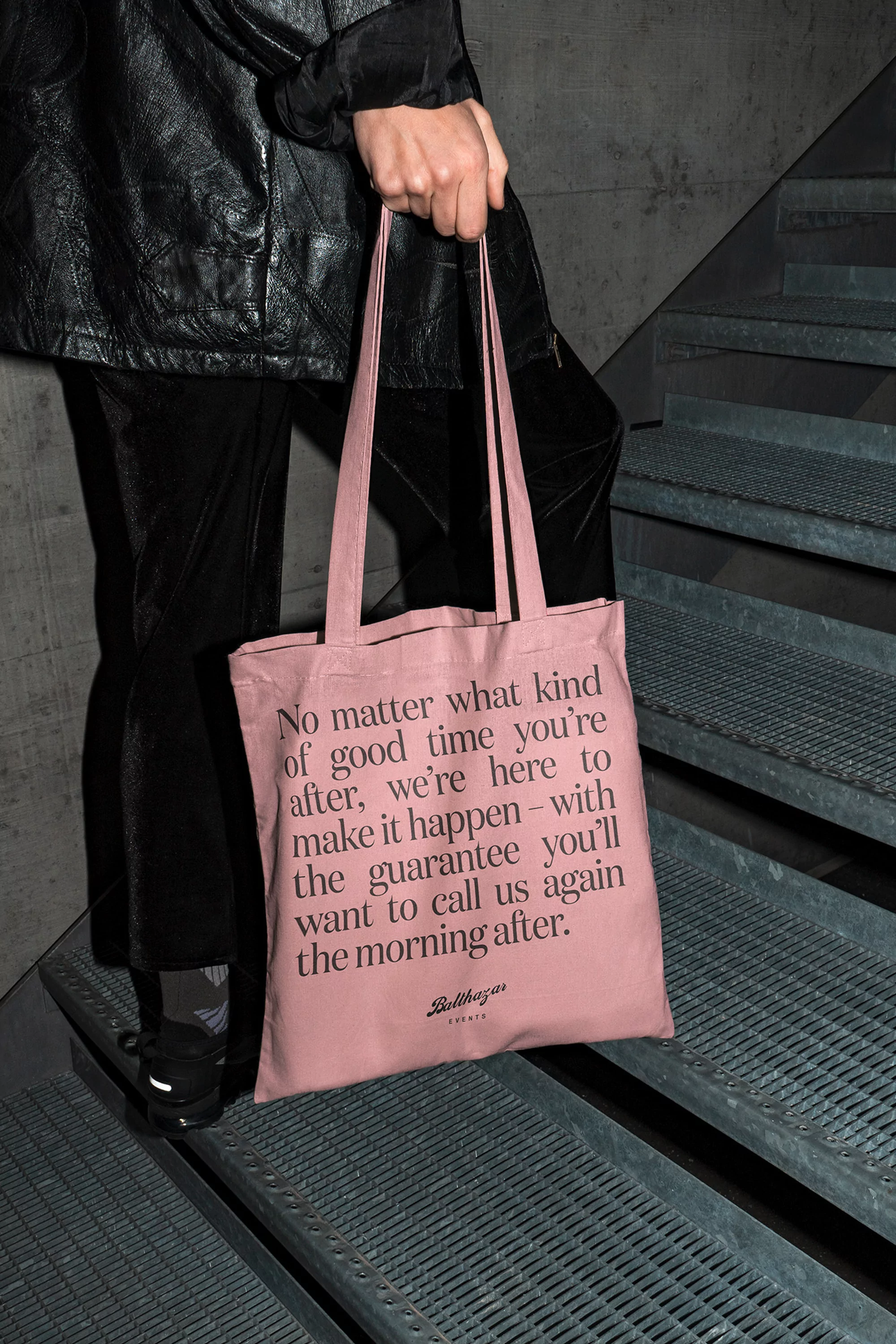 Person holding pink tote bag with printed text on gray stairs, showcasing fashion accessory and unique design against urban backdrop.