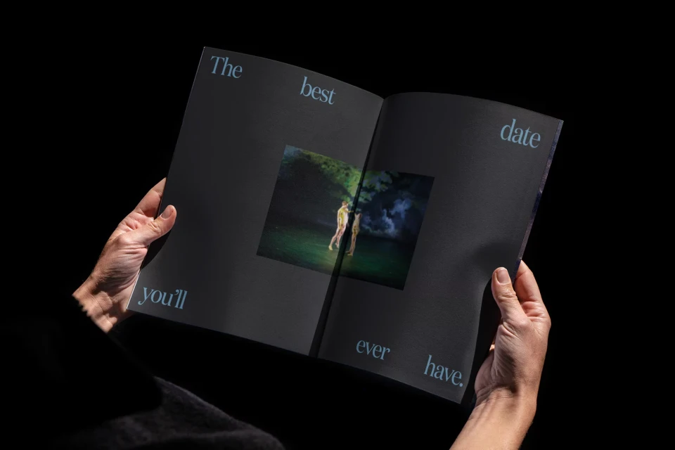 An opened magazine is being held up, featuring an image of two figures in nature, along with text that reads, 'The best date you'll ever have.'