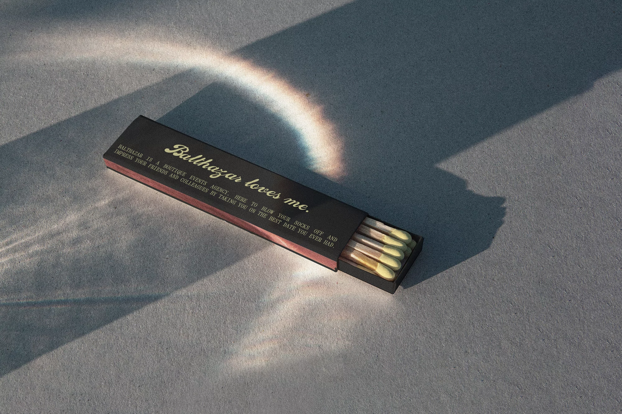 Elevated shot of a black matchbox with yellow-tipped matches, adorned with 'Balthazar loves me' text, set against a grey textured surface.