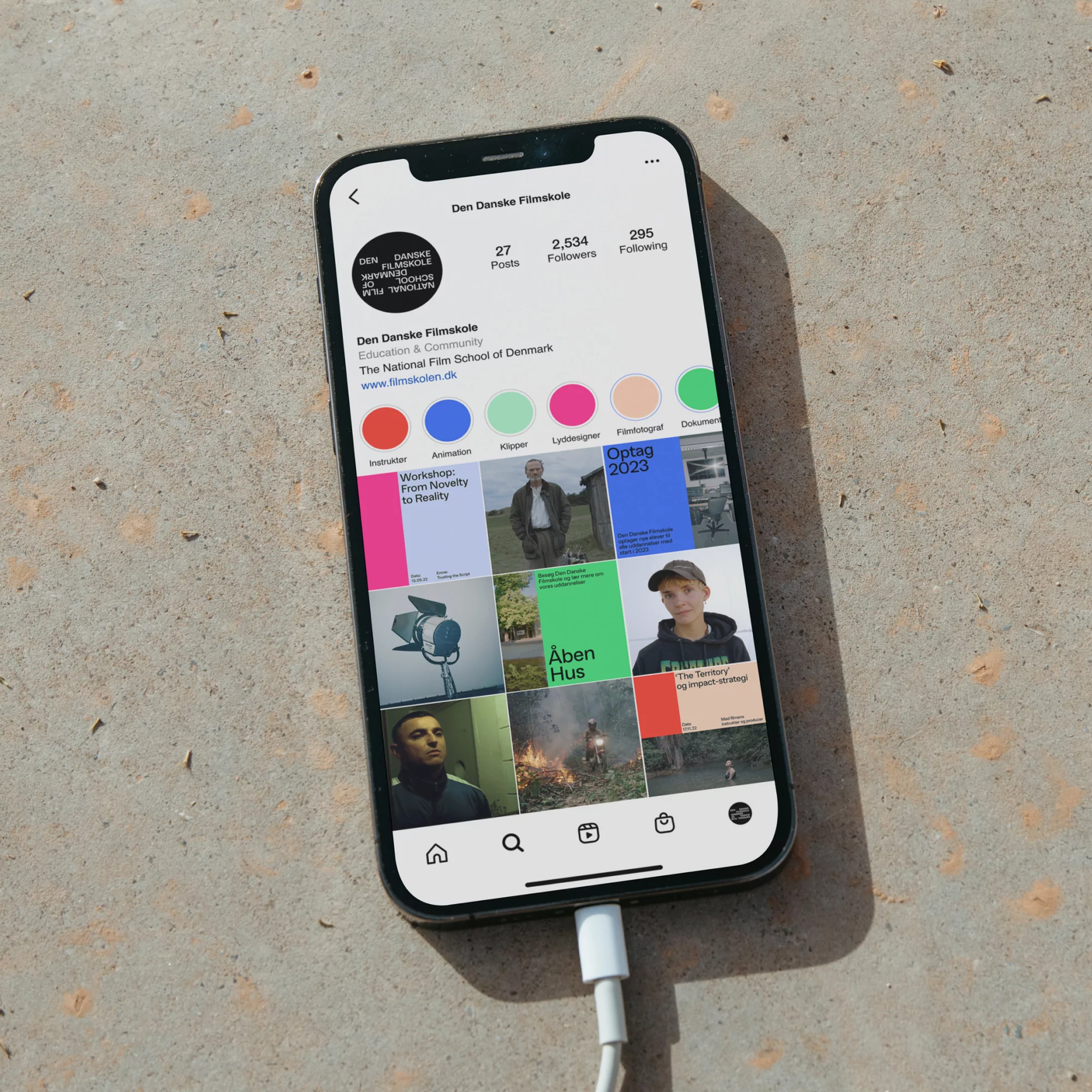 Smartphone displaying the Instagram profile page for the Danish Film School, placed on a concrete surface with its charging cable connected.