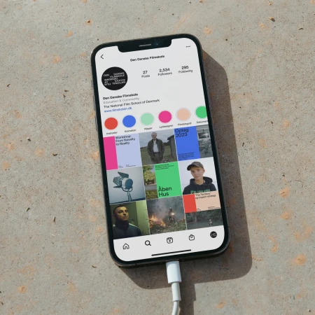 Smartphone displaying the Instagram profile page for the Danish Film School, placed on a concrete surface with its charging cable connected.