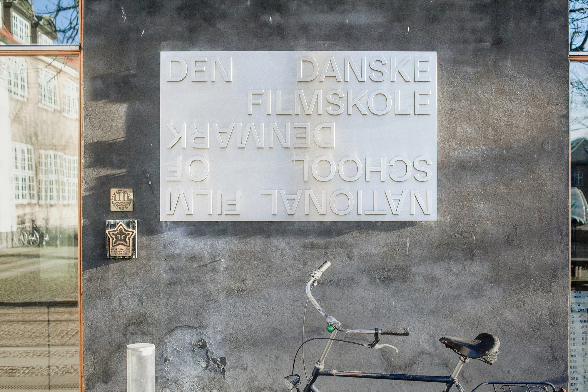 Exterior shot of The National Film School of Denmark sign on a textured grey wall, with a bicycle parked in front of it and a building reflection.