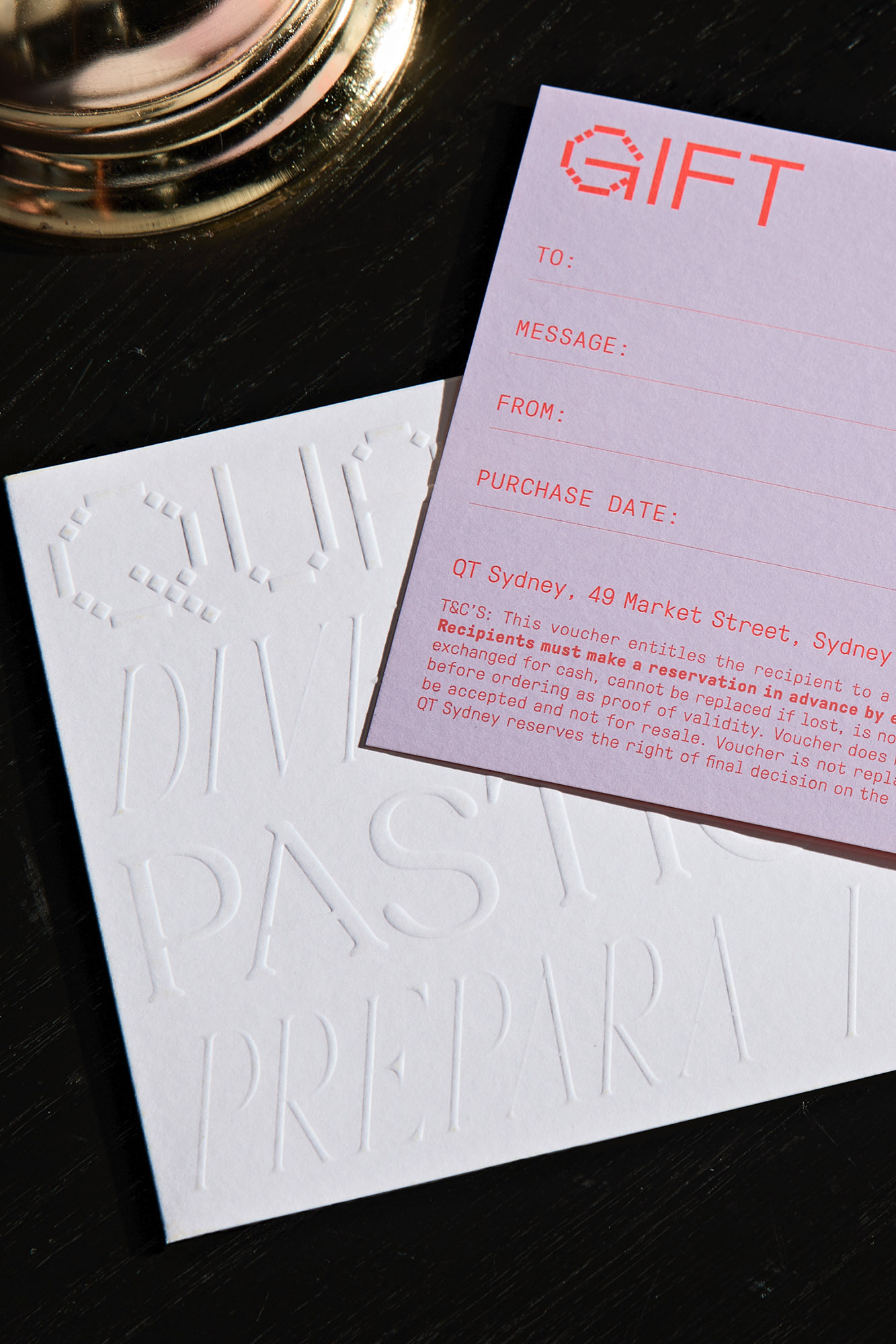 Close-up of a white gift certificate from QT Sydney with embossed lettering and a pink gift voucher on a dark surface. 