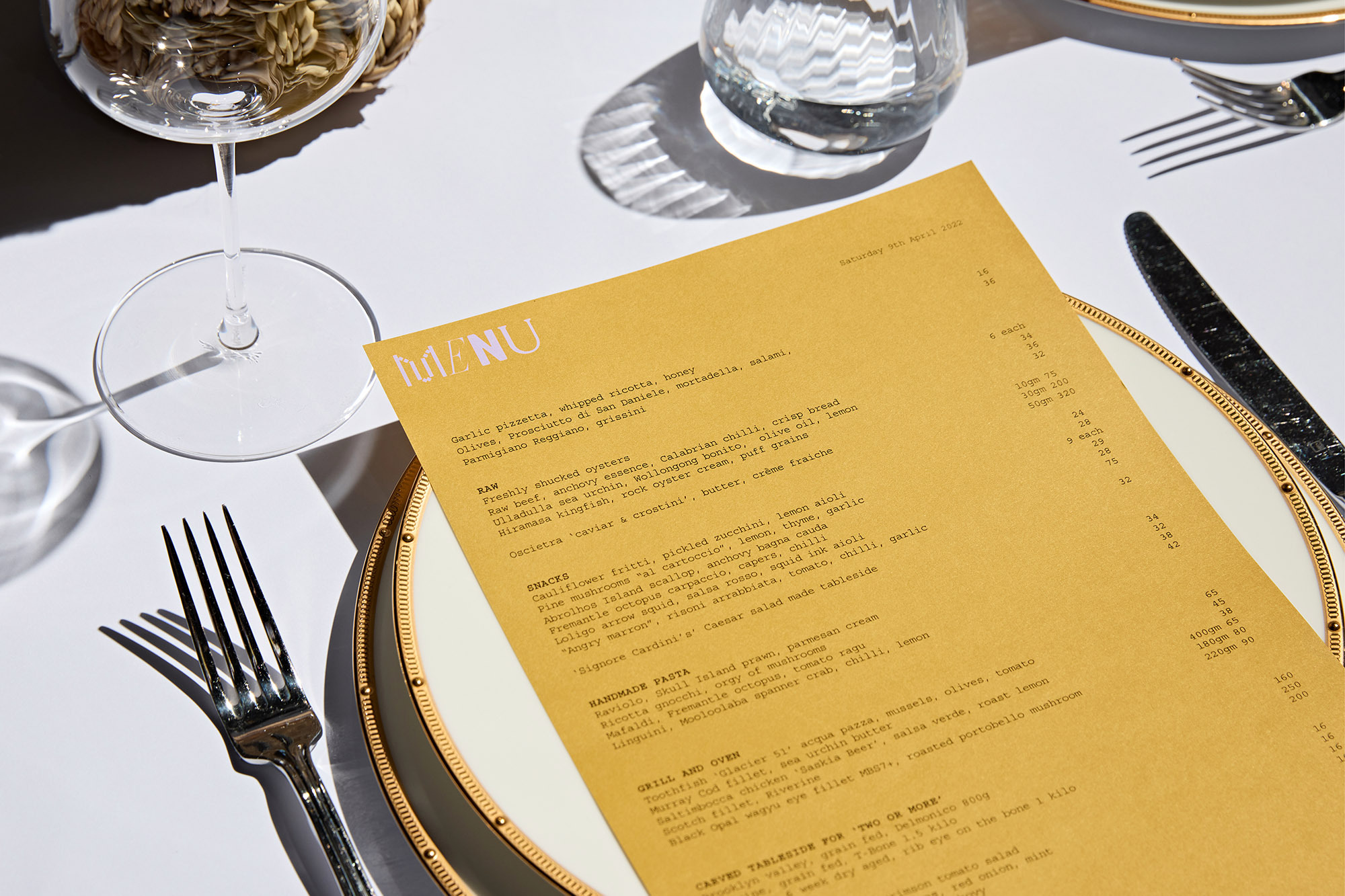 Restaurant menu on white tablecloth with wine glass, silverware, and gold-rimmed plate featuring a golden-toned menu card with text.