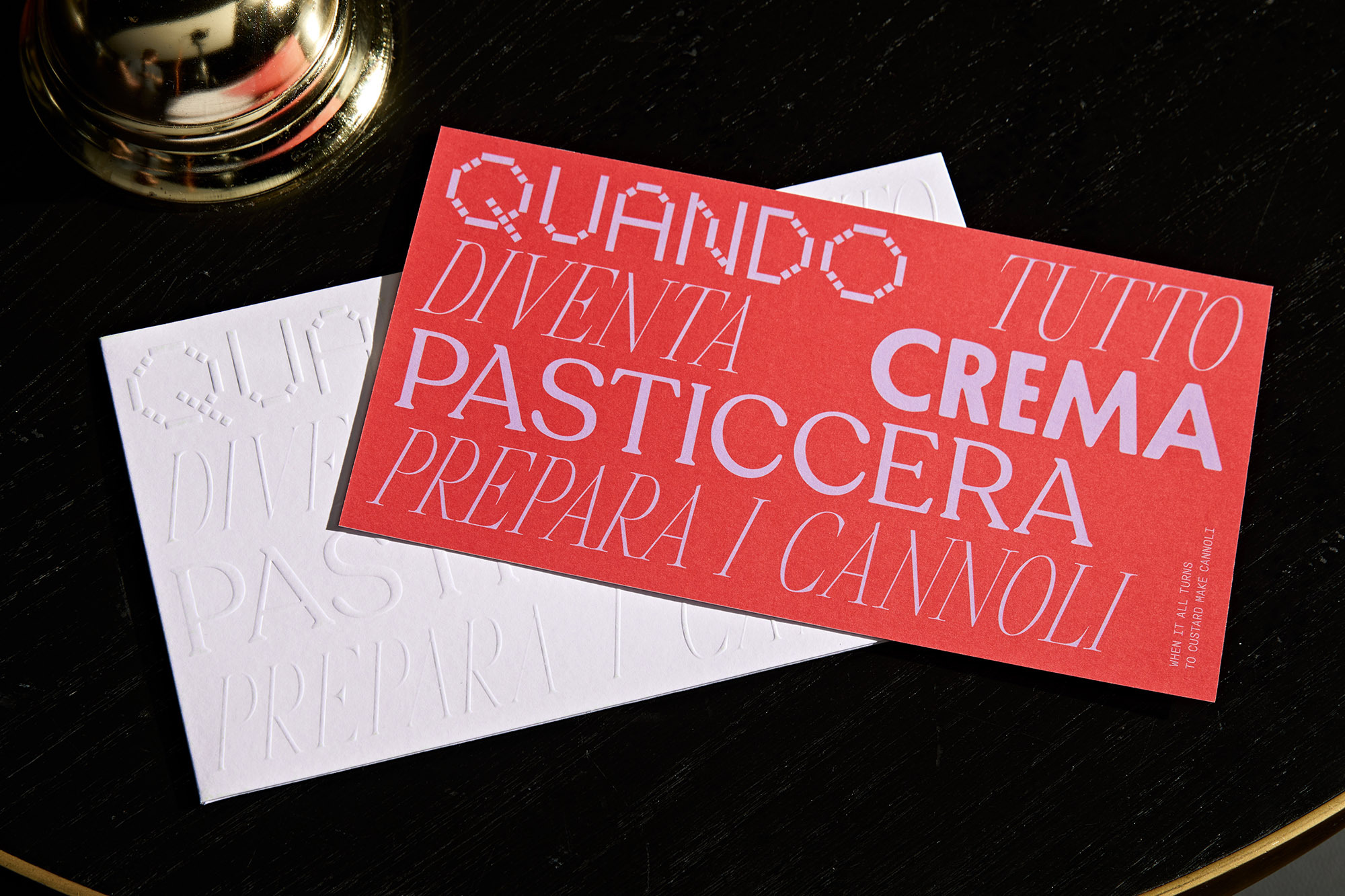 Close-up of two cards on a black surface. One red card features Italian text in lavender. One white card features embossed Italian text.