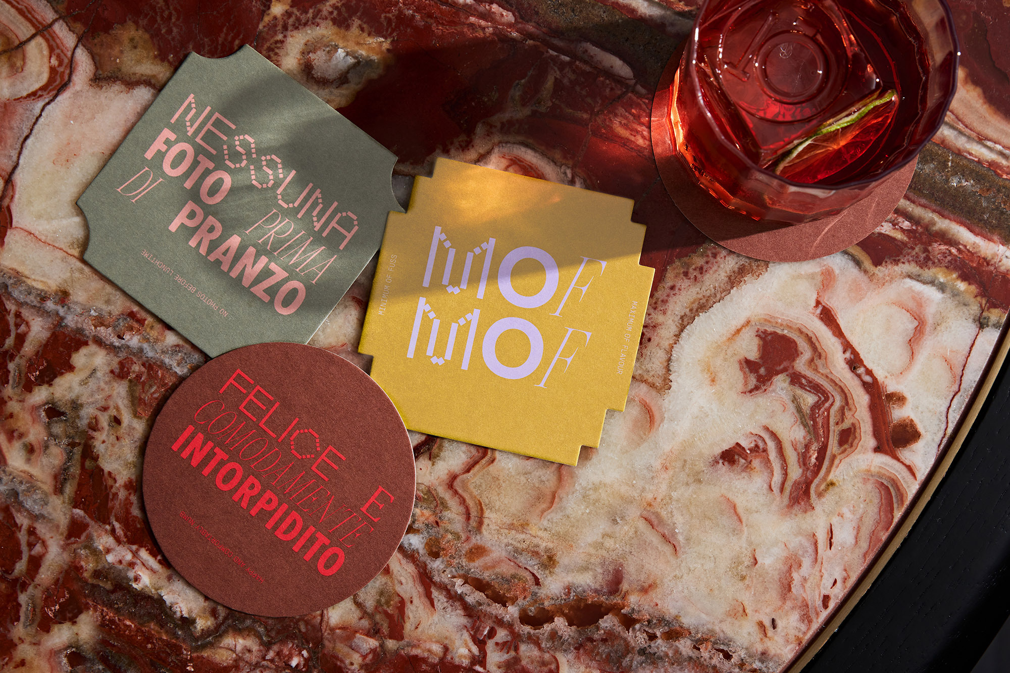Overhead shot of craft cocktail and coasters with Italian text on a marble table with a decorative surface in a warm, inviting setting.