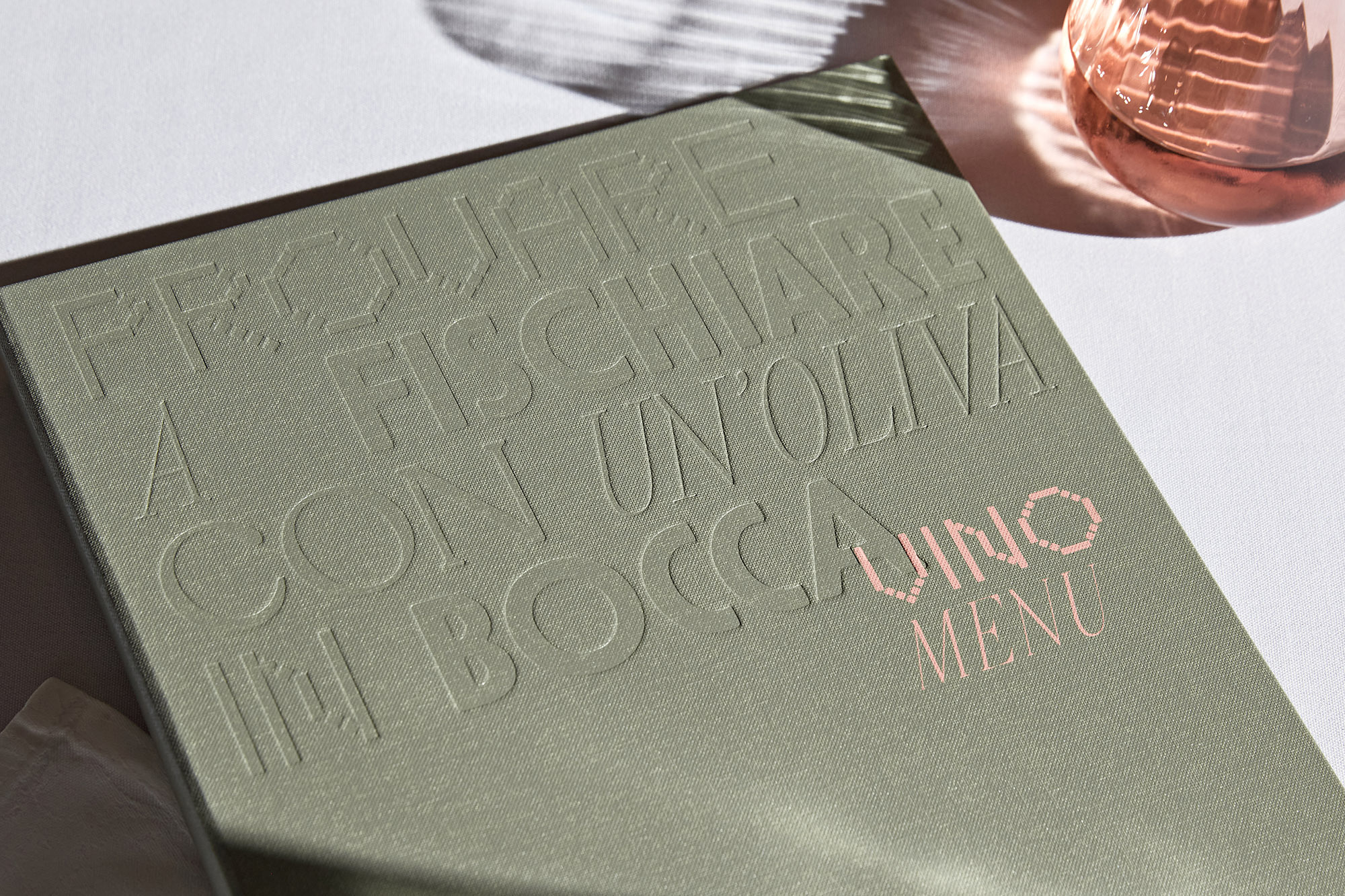 Close-up of a menu cover in olive green with embossed sans-serif typography and pink text. The cover sits on a white surface.