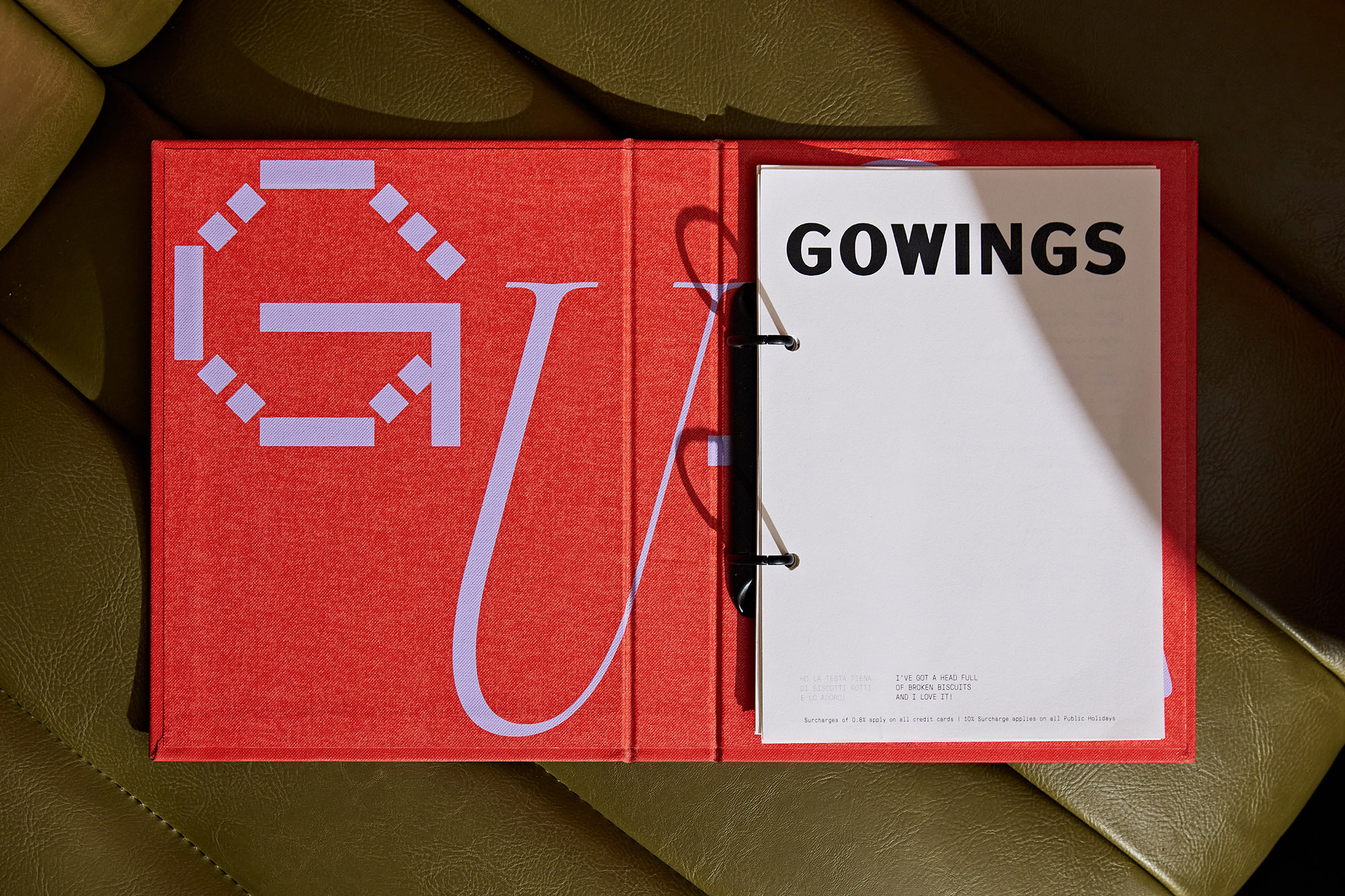 Open coral-colored binder with lavender text and a black ring binder holds white paper labeled 'GOWINGS' on green leather surface.
