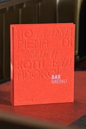 Red textured hardcover bar menu book standing upright on a dark red surface with debossed Italian text and white lettering