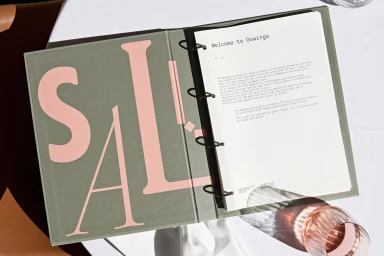An open ring binder menu with sage green cover and pink lettering next to a glass on a table.