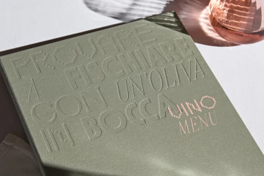Close-up of an olive green menu book with debossed Italian text and a pink glass bottle on white surface