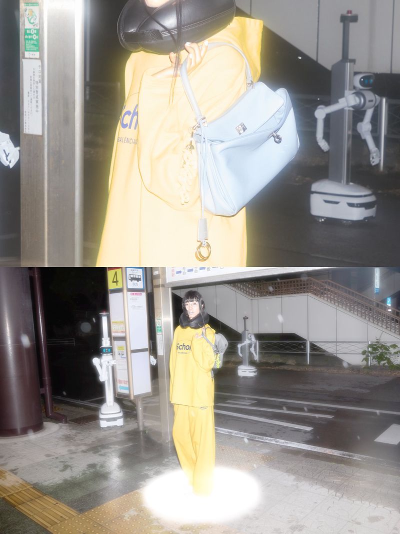 Woman in a yellow outfit carries a blue handbag while standing near a white robot on a city street at night. 
