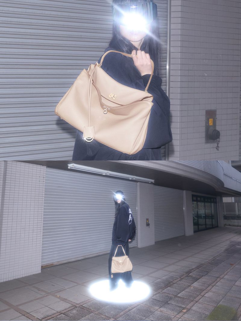 Woman models a beige handbag in an urban setting. She wears a dark outfit in a spotlit area with a concrete wall as a backdrop.