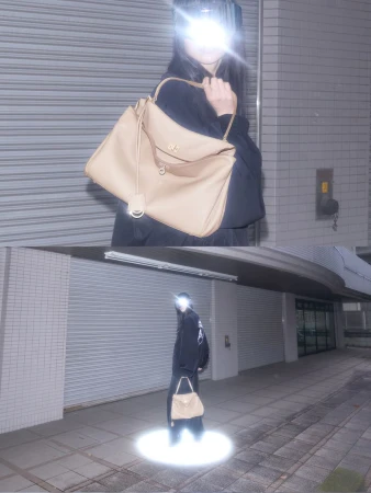 Woman models a beige handbag in an urban setting. She wears a dark outfit in a spotlit area with a concrete wall as a backdrop.