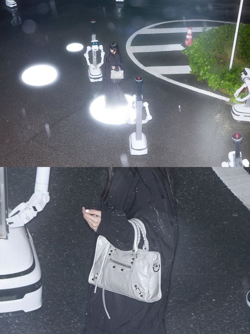 Person in black walks on wet street with service robots, carrying a grey handbag, amidst light reflections and wet asphalt.