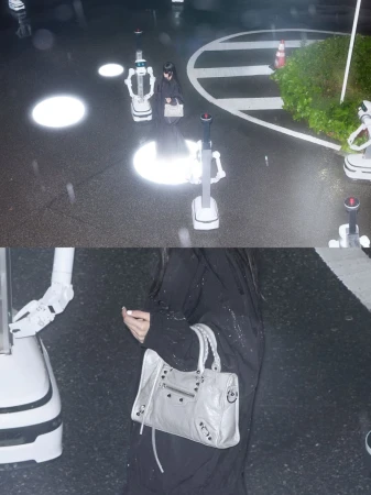 Person in black walks on wet street with service robots, carrying a grey handbag, amidst light reflections and wet asphalt.