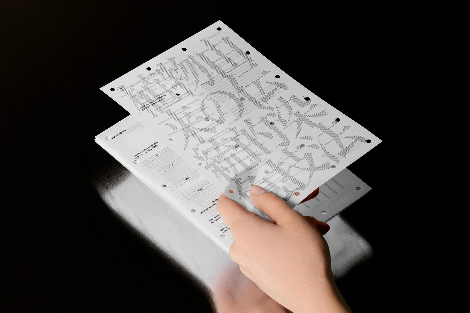 Close-up of hands holding sheets of paper with monochrome typography and graphic elements on a dark reflective surface.