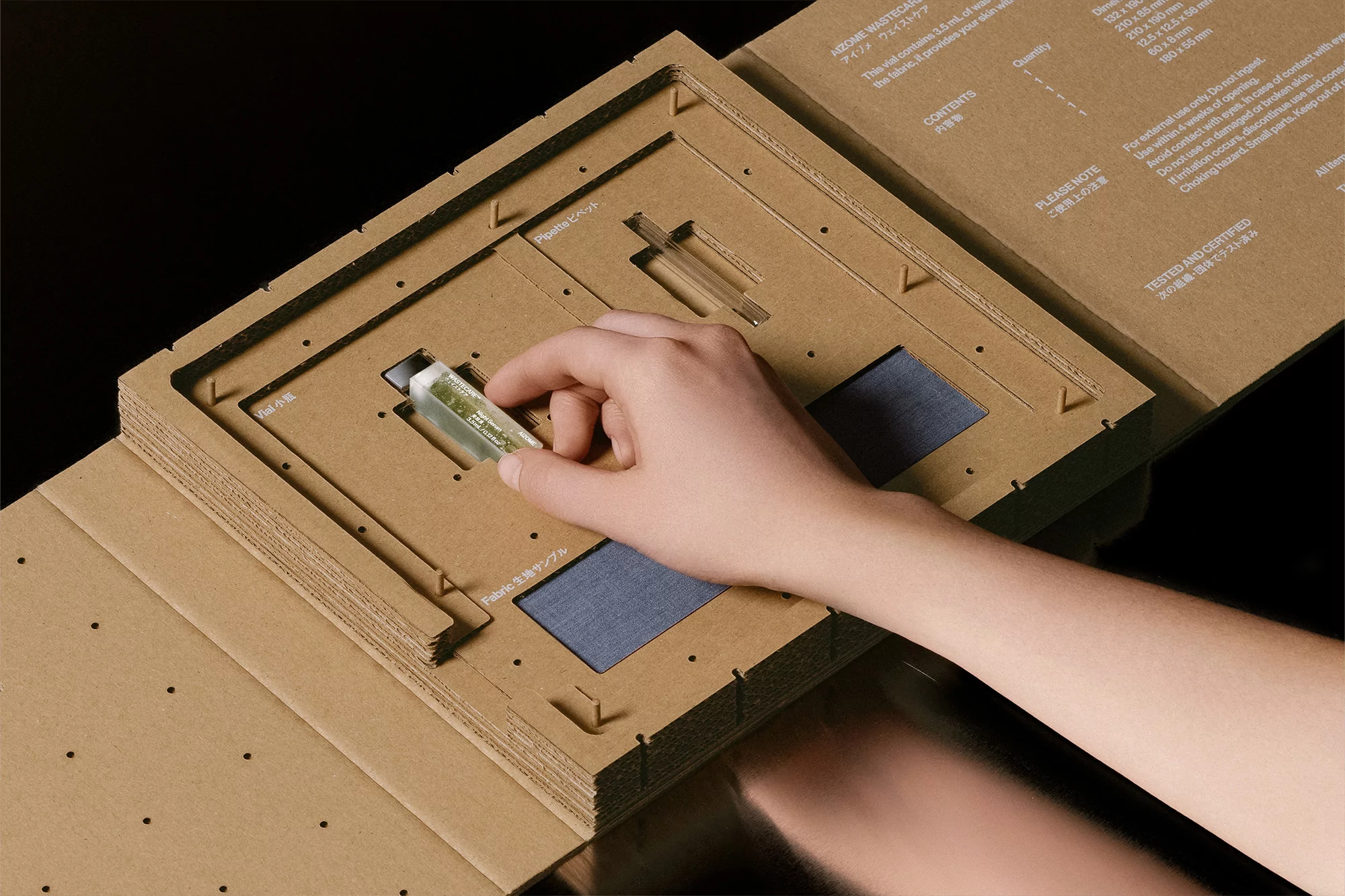 Hand places a small, labeled vial into a corrugated cardboard box with fabric samples against a black background in a studio setting.