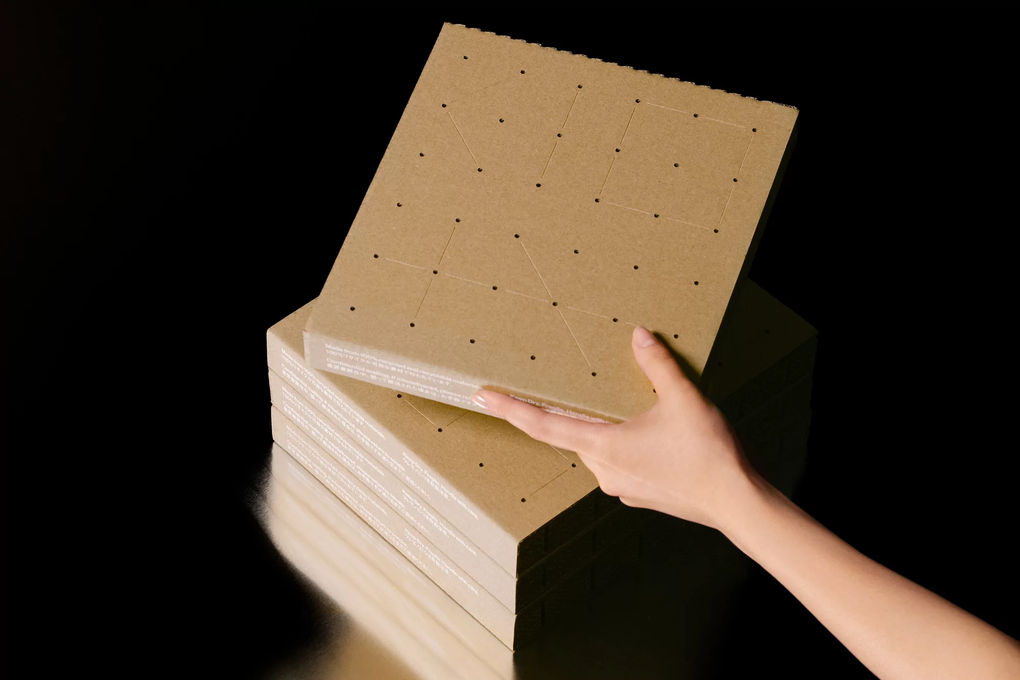 A hand holding a square tan cardboard sheet with a geometric pattern of dotted lines, atop a stack of similar sheets against black.