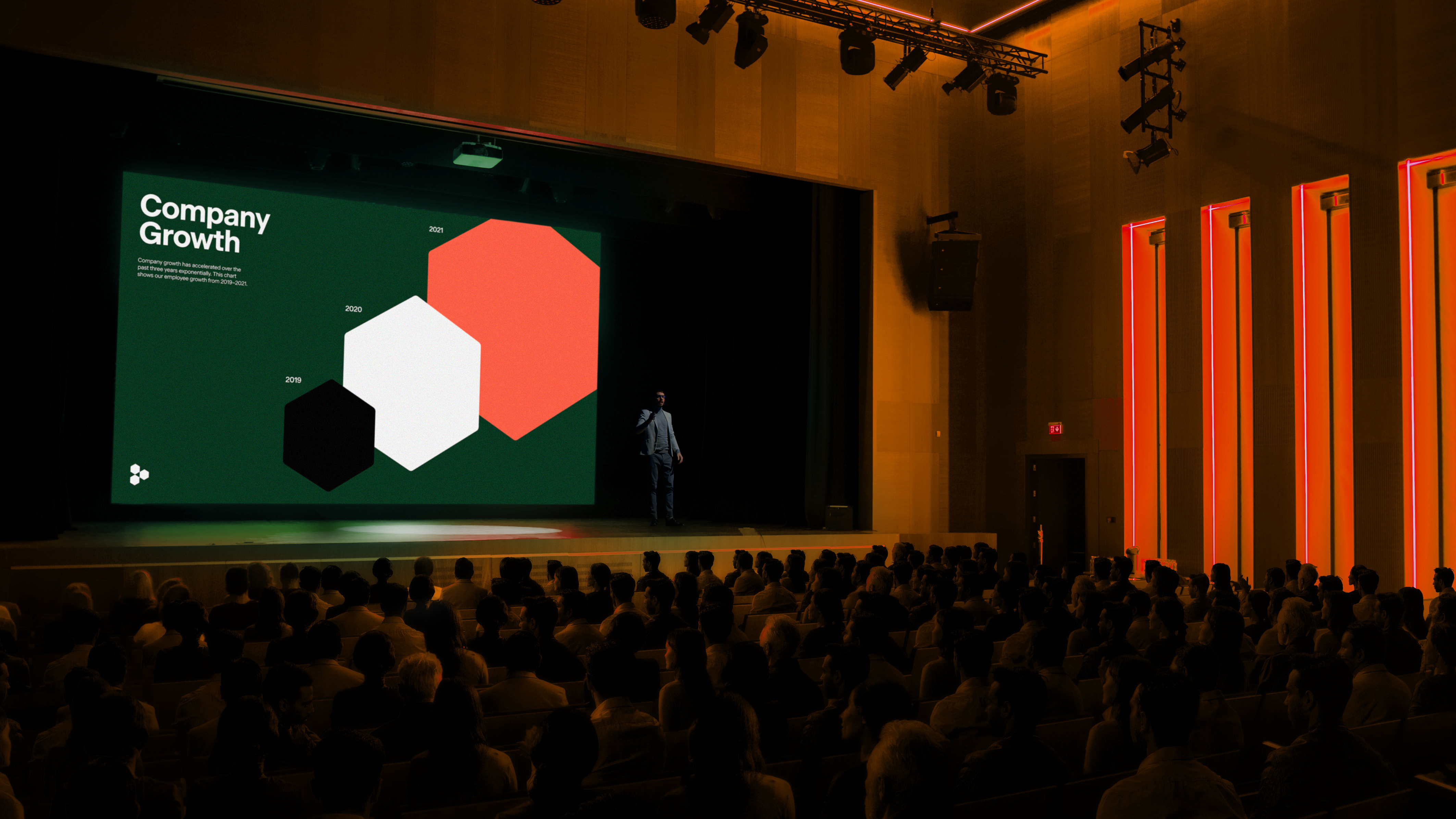 Audience views presenter on stage delivering business presentation with company growth graphs and color-blocked hexagons on screen.