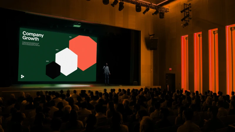 Audience views presenter on stage delivering business presentation with company growth graphs and color-blocked hexagons on screen.
