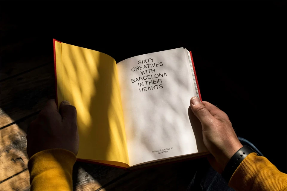 Open book with title 'Sixty Creatives with Barcelona in Their Hearts', held by two hands, warm light illuminating the pages.