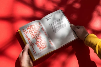 Open book featuring red typography held by hands wearing a yellow sleeve against a vibrant red background with leaf shadow patterns.