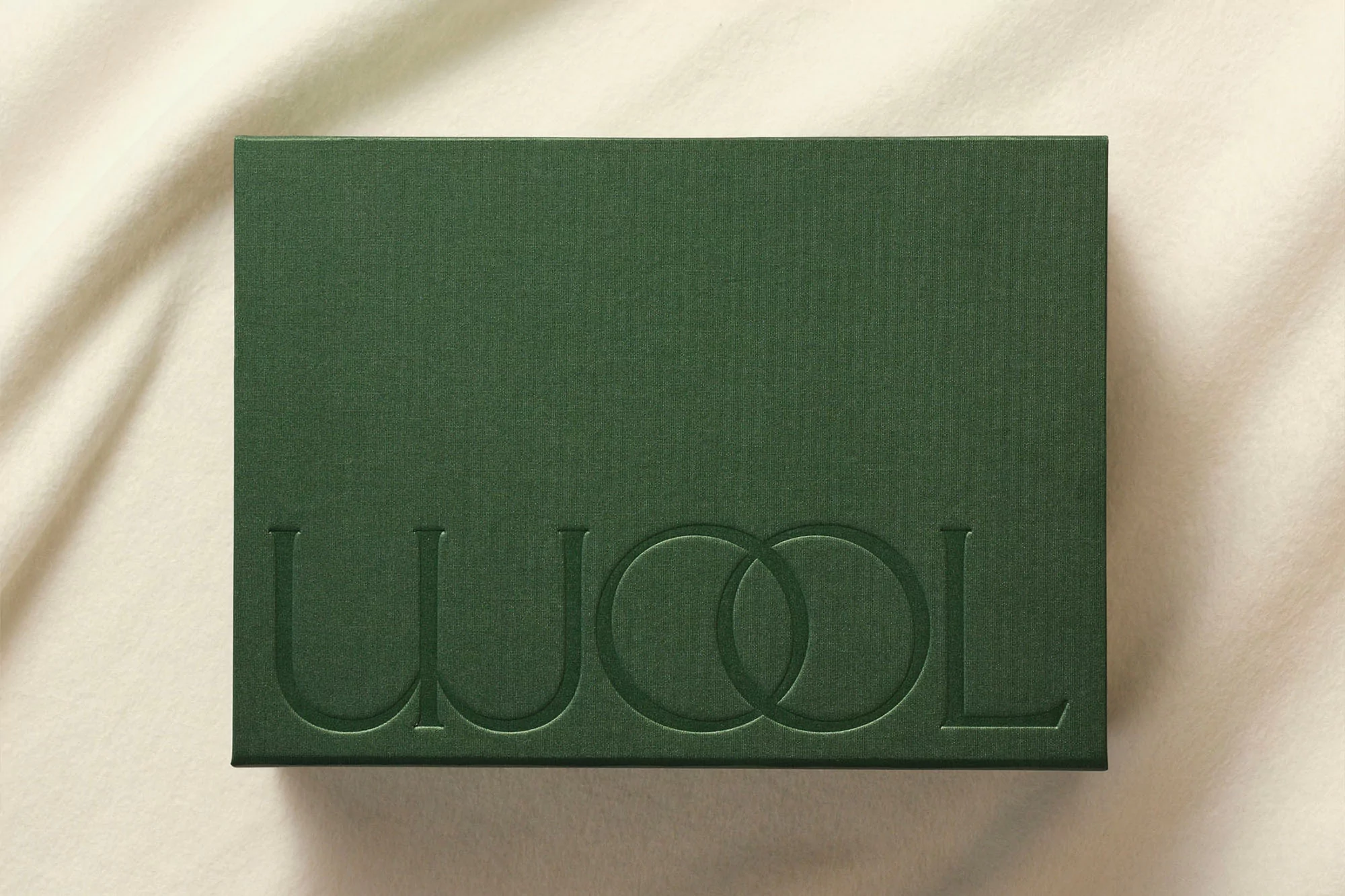 Dark green box featuring the word 'WOOL' in debossed capital letters, resting on a softly draped, light cream-colored fabric backdrop.