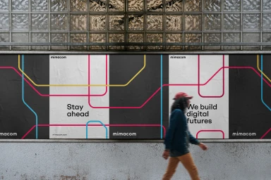 A person walking past a large billboard poster displaying the Mimacom brand identity with graphic transit map lines.