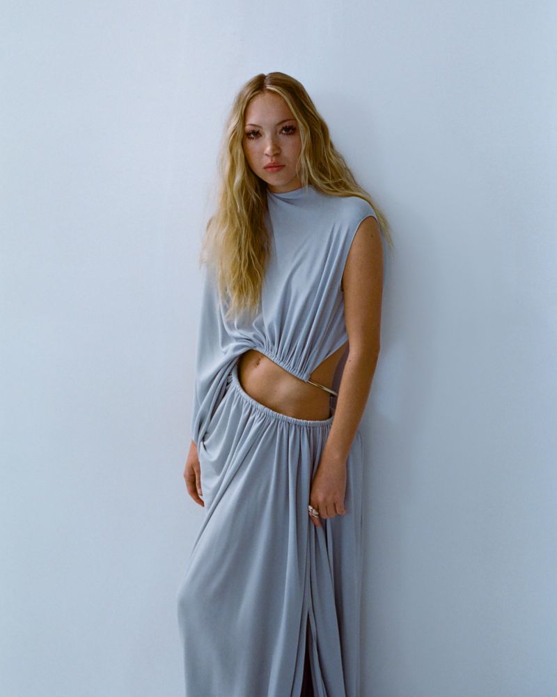 A young woman with long blond hair poses in a grey, flowing gown featuring cut-out details, set against a light-grey background.