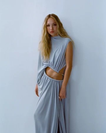 A young woman with long blond hair poses in a grey, flowing gown featuring cut-out details, set against a light-grey background.