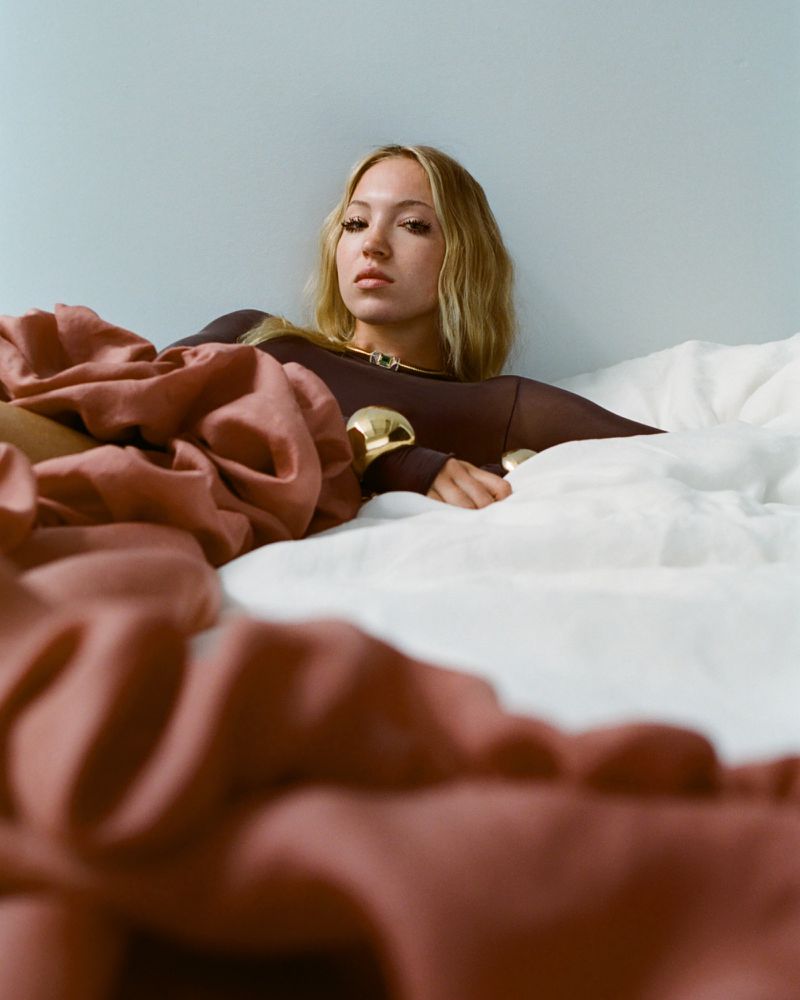 Blonde adult female with sheer brown top and gold jewelry lying on white sheets with red draped fabric in a studio setting.