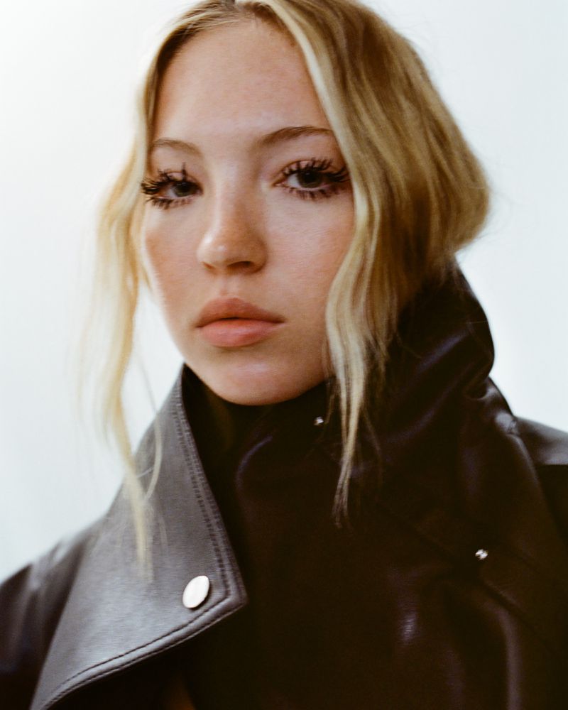 Close-up portrait of a young adult woman with blonde hair wearing a black leather jacket against a plain white background.
