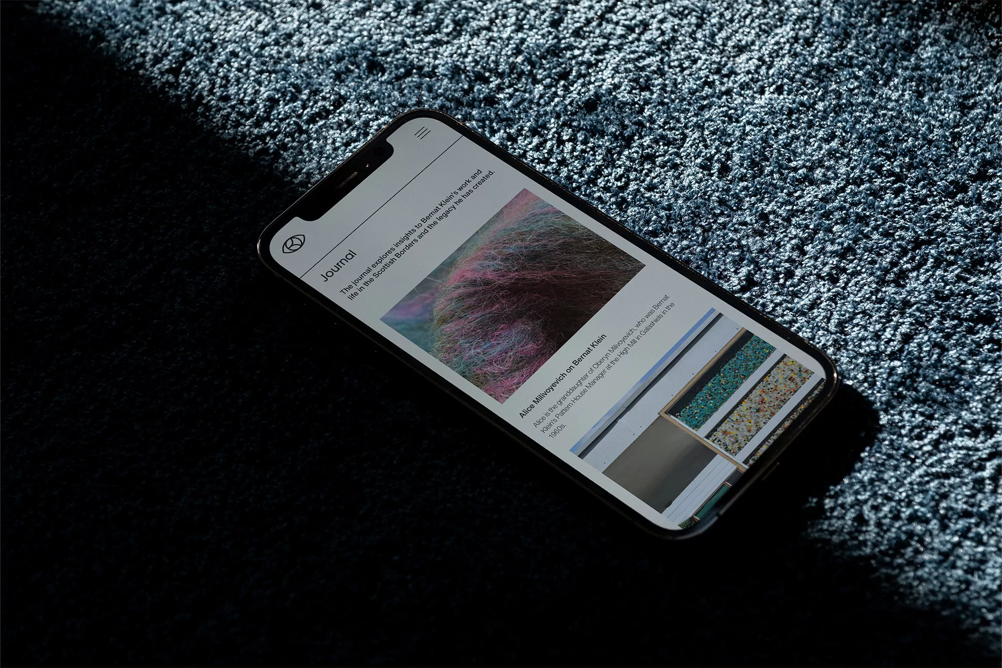 Black smartphone displays a Journal app webpage with design articles, resting on a plush textured blue carpet under soft, directional light.