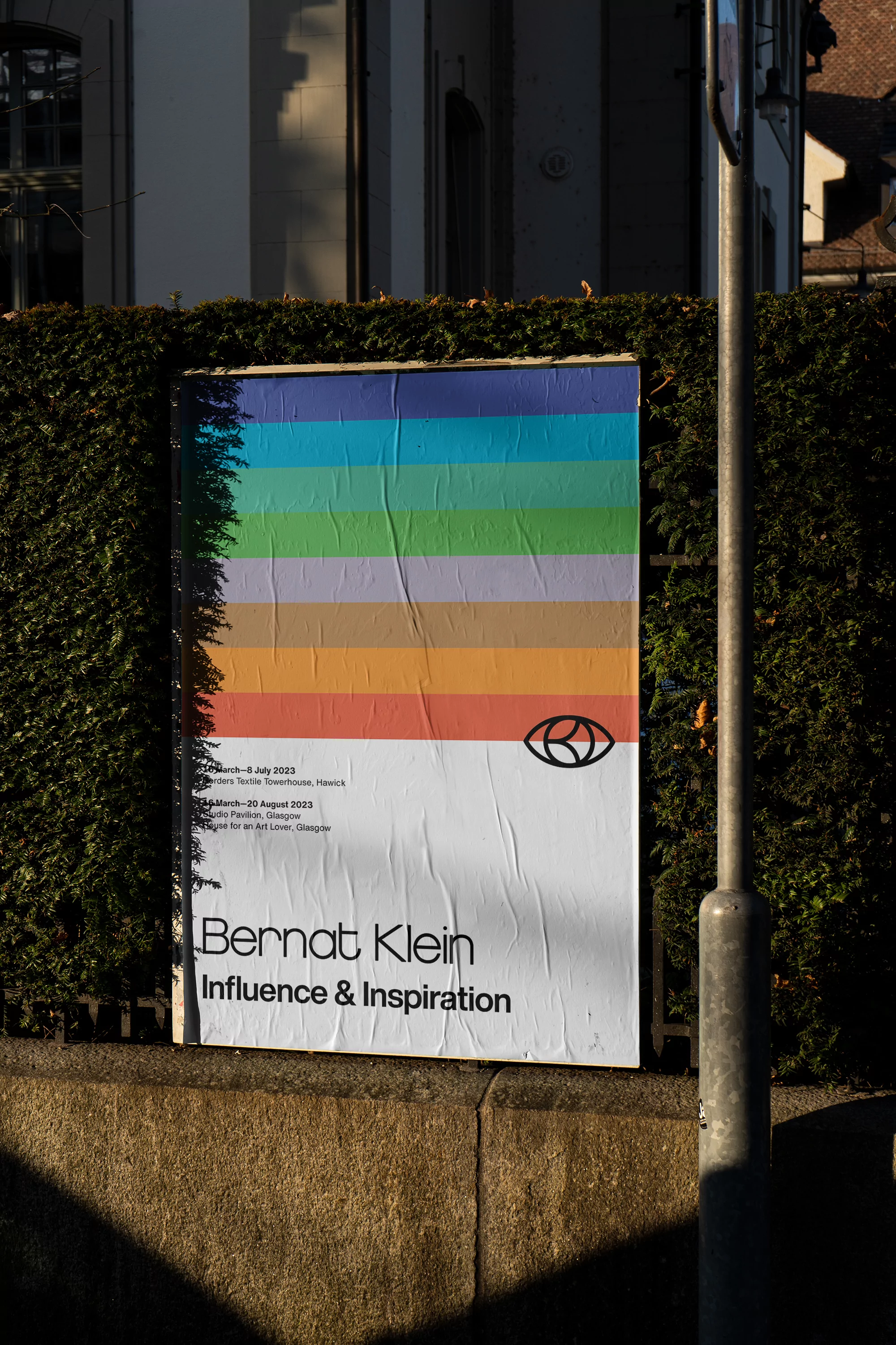 A colorful poster featuring rainbow stripes and text is mounted on a hedge-covered wall beside a streetlight in an outdoor setting.