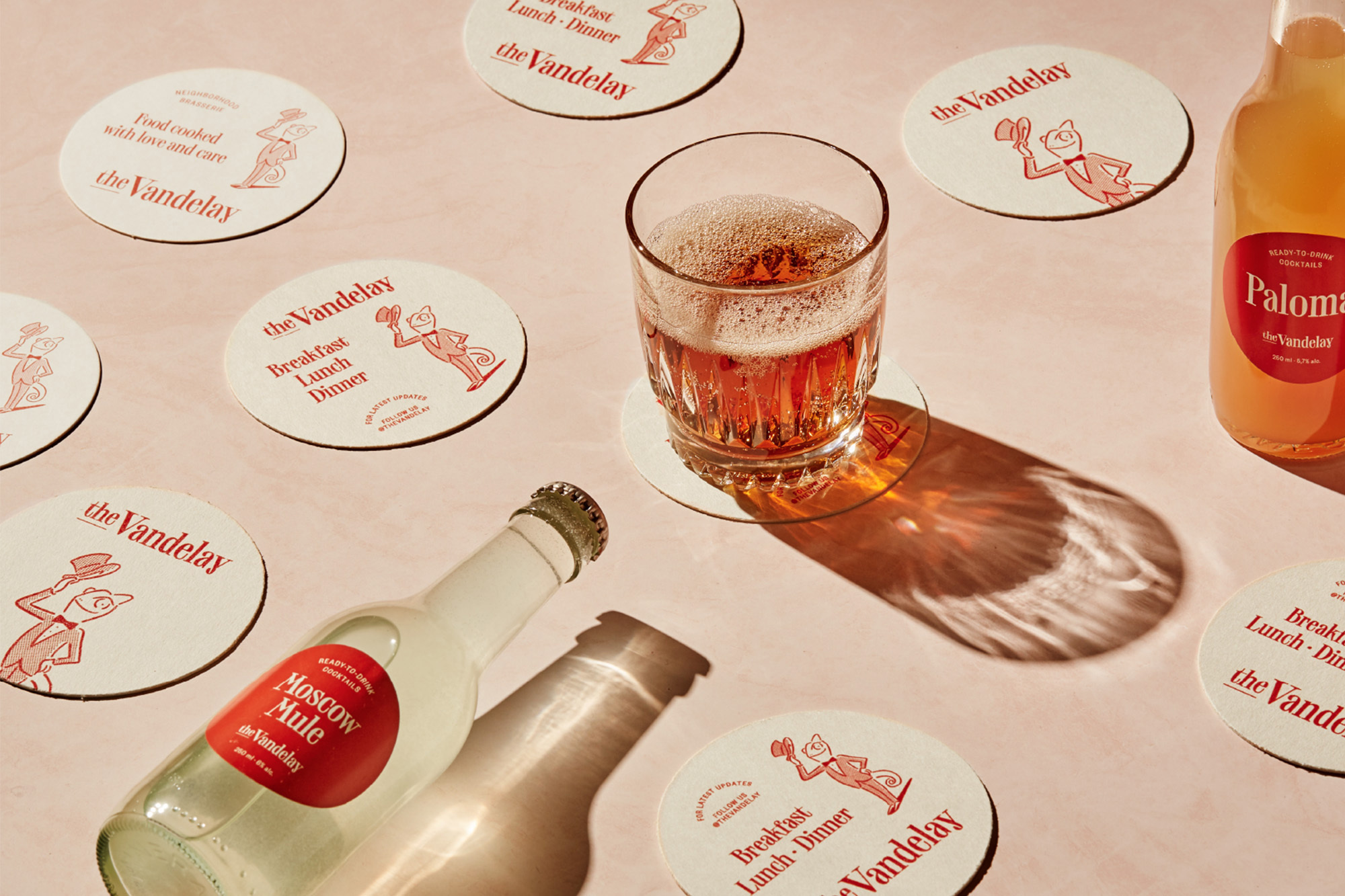 Overhead shot of a cocktail in a glass, surrounded by branded coasters and bottles on a pale pink surface in soft lighting.