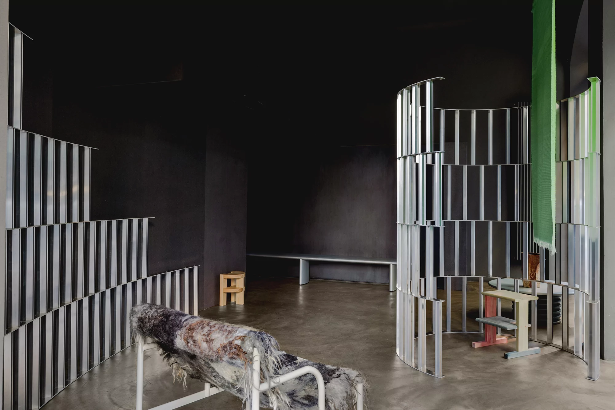 Abstract interior scene features gray concrete floor, black walls, decorative metal screens, and textile-draped chaise lounge in a studio setting.