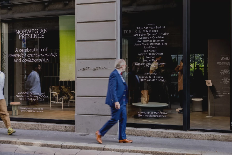 Man in blue suit walking past storefront window displaying 'Norwegian Presence' exhibition details, featuring furniture.