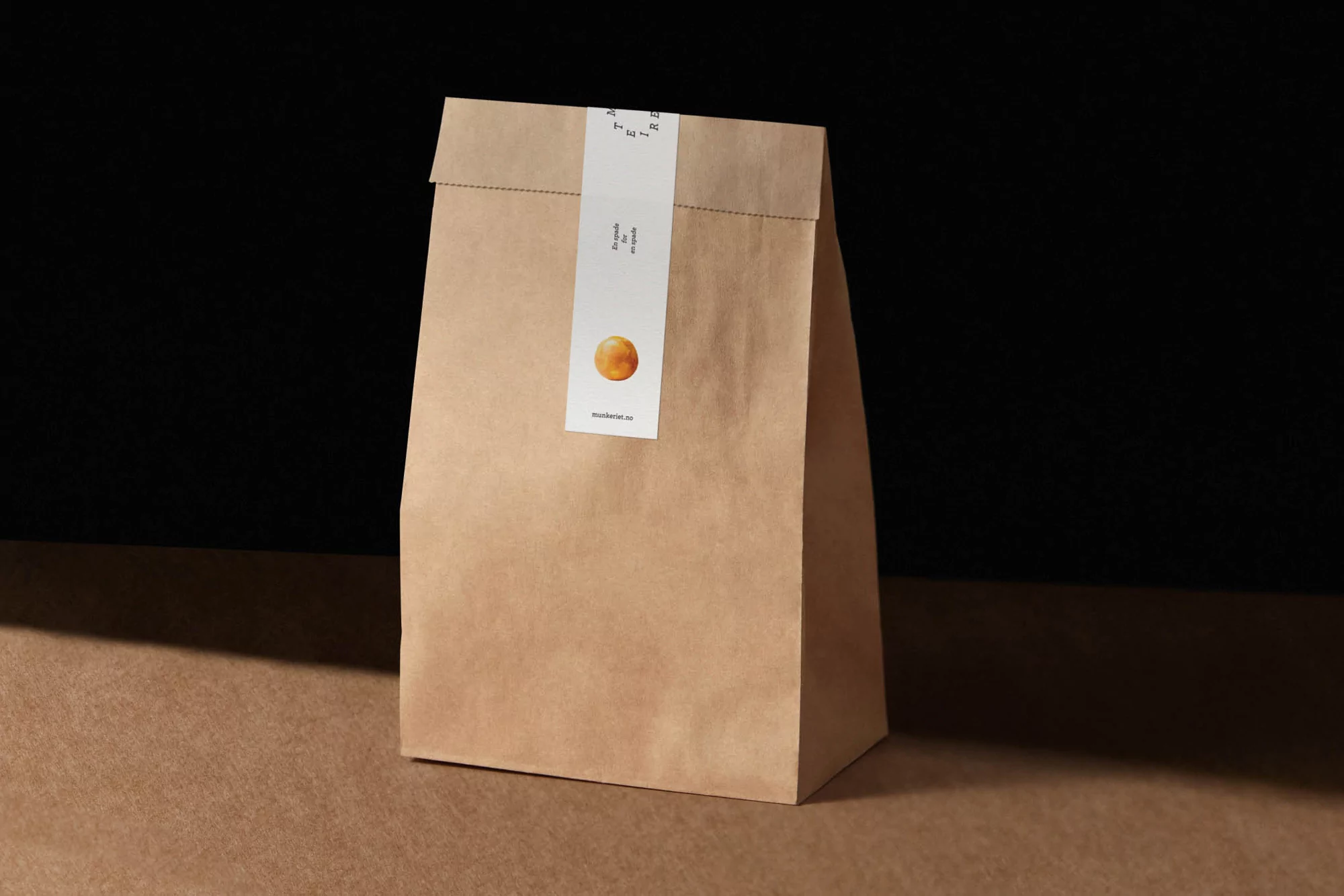 Studio shot of a kraft paper bag sealed with a label featuring a round orange graphic, standing on a kraft paper surface against a black background.