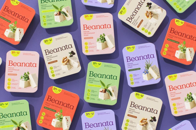 Overhead shot of colorful Beanata vegan cheese packaging, displaying cubes of cheese with various toppings, arranged on a purple surface.