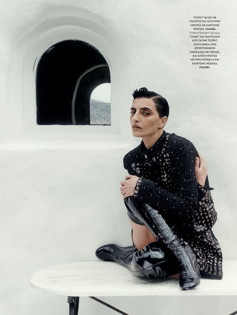 Woman with short hair wearing black dress and black shiny boots poses sitting on white table in front of whitewashed wall.