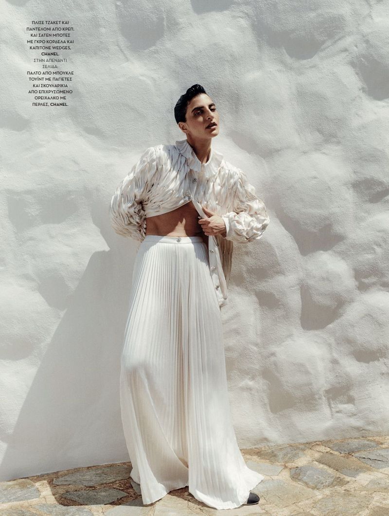 An adult female model poses against a white wall wearing a ruffled jacket, wide-leg pleated trousers, and dark shoes in an outdoor setting.
