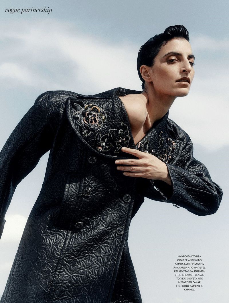 Fashion editorial shot of a woman in a black patterned coat against a sky background. Vogue partnership advertisement.